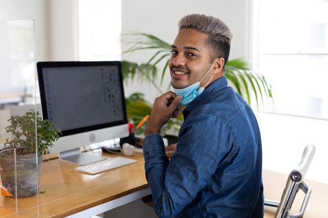 A caucasian man sitting on his desk in front of the computer at the office. he is pulling his facemask down revealing a smile.