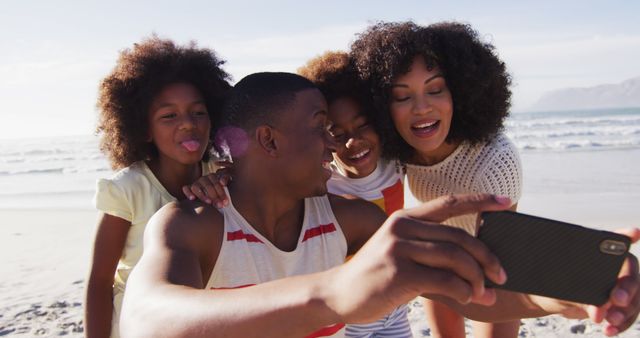 Happy African American family with parents and children enjoying time together at the beach, capturing joyful moments with a smartphone selfie. Perfect for themes related to family bonding, vacations, summer, and healthy relationships.