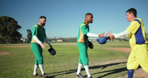 Diverse baseball teammates exchanging caps and shaking hands walking on sunny field