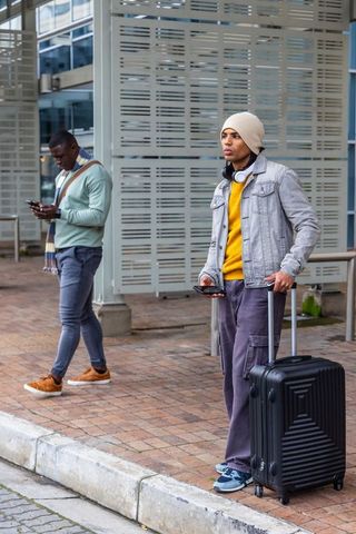 Young male travelers standing curbside with suitcase and phones in urban transit wearing beanie