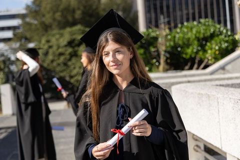 Smiling woman graduating in black gown and mortarboard holding diploma on sunlit campus
