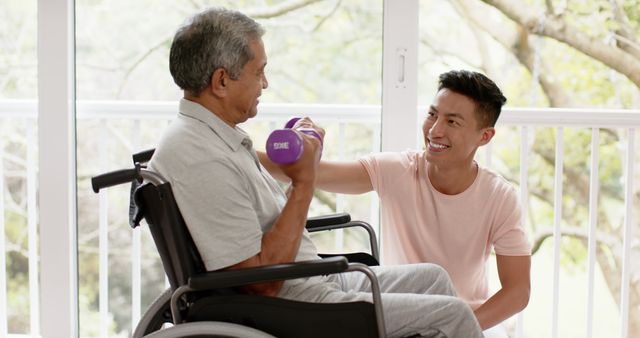 Senior man in a wheelchair lifting dumbbell with help from young caregiver indoors. Ideal for use in articles and advertisements related to elderly care, fitness programs, rehabilitation services, and healthcare support. This depiction can also highlight the importance of physical activity and personal assistance for seniors.