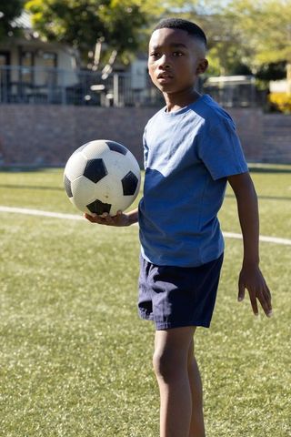 African american boy holding soccer ball on sunlit turf wearing blue shirt and navy shorts