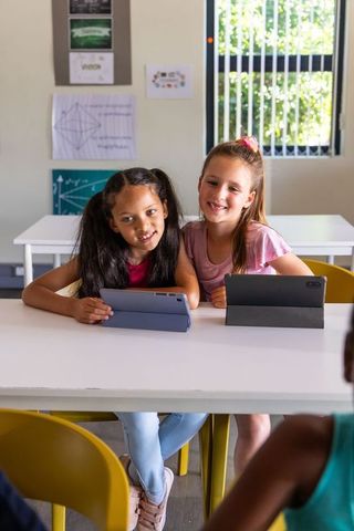 School-age girls collaborating on tablets in bright classroom with greenery view