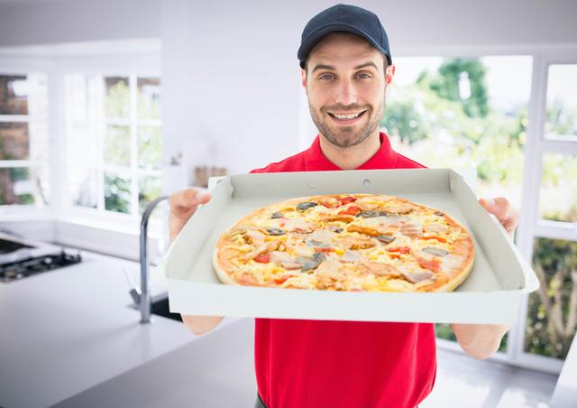 Smiling deliveryman holding a pizza box in a modern kitchen. Ideal for use in advertisements for food delivery services, restaurant promotions, or articles about convenience and fast food. Can also be used in marketing materials for culinary services or home meal delivery.