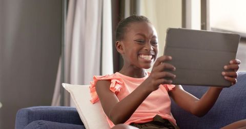 African american girl laughing and playing on tablet at home on cozy couch in sunlight