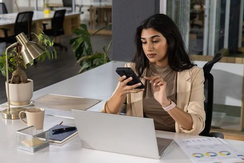 Indian businesswoman checking smartphone at open-plan office desk with laptop and charts