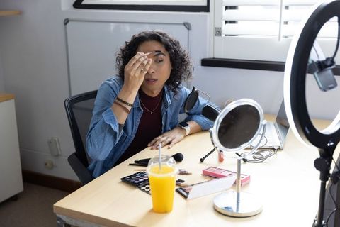 Non-binary creator applying makeup at home workstation with ring light and vanity mirror
