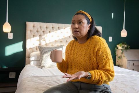 Mature woman sitting on bed holding ceramic mug and gesturing in cozy green bedroom