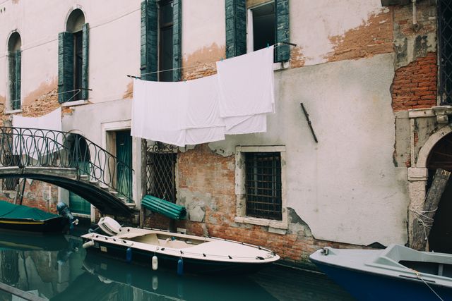 This image showcases clothes drying above a Venetian canal, with boats docked near a rustic historic building. Ideal for travel blogs, articles on Venice, photography inspiration, or European architectural studies.