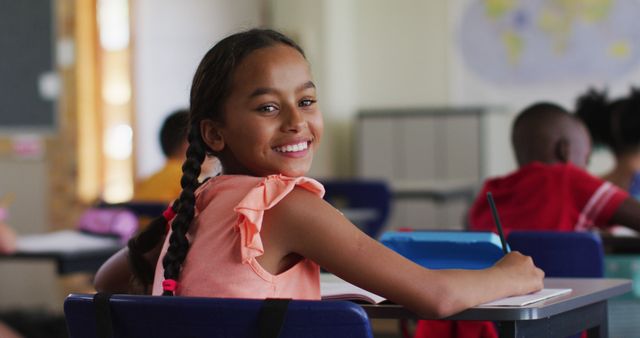 Portrait of happy biracial schoolgirl sitting at classroom, making notes, looking at camera. children at primary school