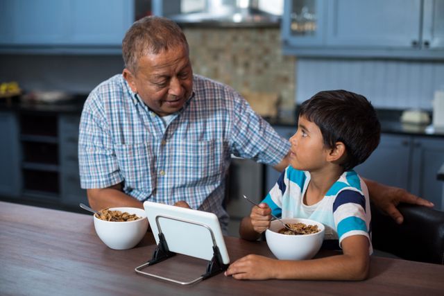 Grandfather and grandson sitting at kitchen table, eating cereal and using tablet. Ideal for themes of family bonding, technology in daily life, multigenerational relationships, and morning routines.
