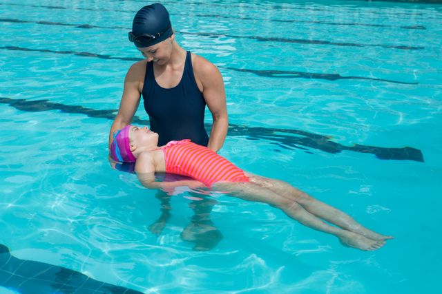 Female swimming instructor training a young girl in a pool at a leisure center. The instructor is wearing a swim cap and holding the girl who is learning to float. Great for content related to swimming lessons, water safety, aquatic skills, and child education.