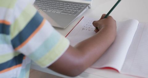 Child writing math homework in lined notebook while using laptop for focused study