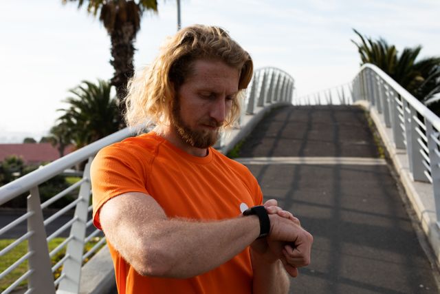 Front view of a fit Caucasian man with long blonde hair wearing sportswear exercising outdoors in the city on a sunny day with blue sky, looking at smartwatch standing on footbridge.