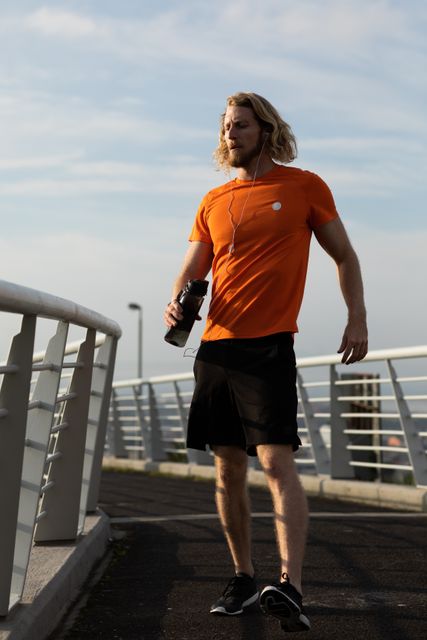 Front view of a fit Caucasian man with long blonde hair wearing sportswear exercising outdoors in the city on a sunny day with blue sky, walking on a footbridge, holding water bottle in hand, wearing headphones.
