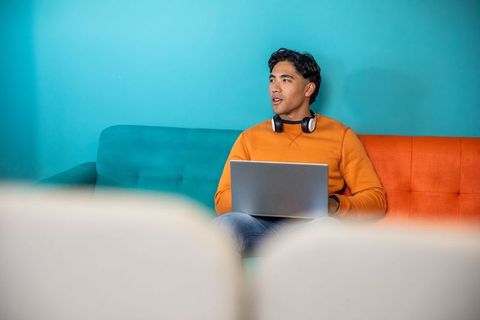Young asian man working remotely with laptop and headphones on vibrant two-tone sofa