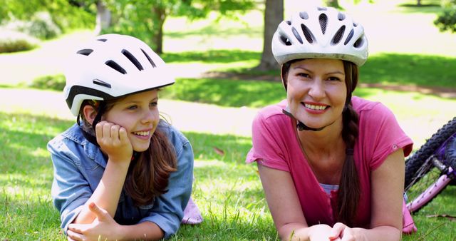 Mother and daughter lying on grass, both wearing bicycle helmets and smiling at camera. Perfect for depicting family bonding, outdoor activities, summer leisure, cycling safety, and healthy lifestyles.