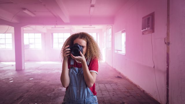 Front view of a hip young Caucasian woman in an empty warehouse, standing and taking photos with SLR camera, wearing a red shirt.