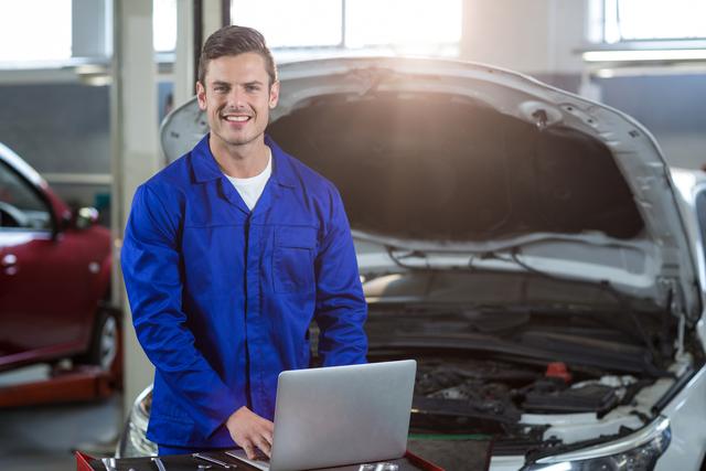Mechanic in blue overalls using laptop for car diagnostics in a repair garage. Ideal for content about automotive technology, car maintenance, professional mechanics, and modern repair techniques.