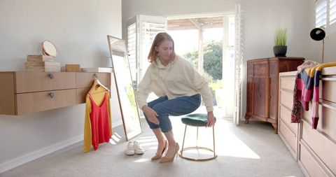 Young woman sitting on stool putting on beige heels in sunlit dressing room casual style