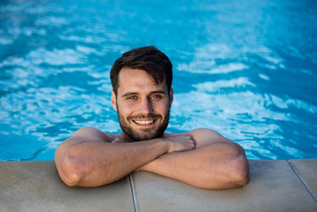 Young man enjoying a sunny day in the swimming pool, leaning on the edge with a cheerful smile. Perfect for promoting summer activities, vacation destinations, leisure time, and lifestyle content.