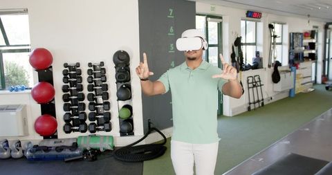a man using a virtual reality headset participates in therapy exercises within a rehabilitation center. surrounding equipment includes medicine balls and dumbbells, emphasizing an active, wellness-oriented environment. this could be utilized to highlight advancements in medical recovery methods, innovative healthcare solutions, or virtual reality applications in therapy.