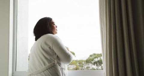 African American woman standing at window in white bathrobe reflecting in morning light