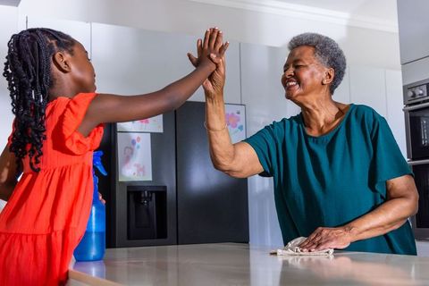 African american grandmother and granddaughter high-fiving while cleaning bright kitchen