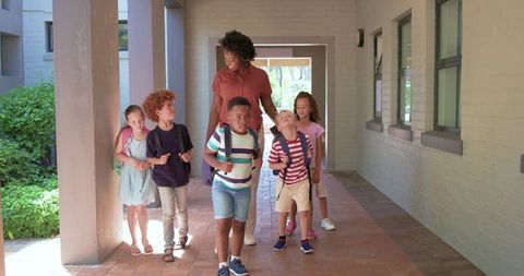Teacher leading diverse elementary students with backpacks walking down brick corridor
