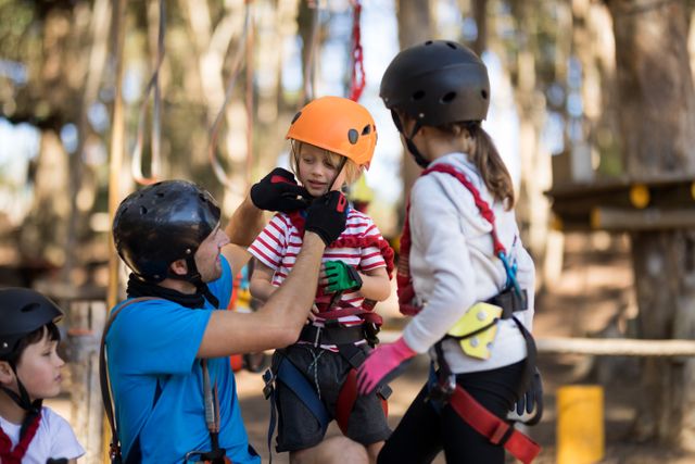 Instructor assisting child with safety helmet in adventure park. Ideal for promoting outdoor activities, family bonding, safety measures, and adventure sports. Useful for educational materials, advertisements for adventure parks, and articles on child safety.