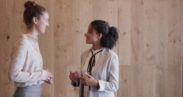 The image shows two professional women engaging in a discussion in an office setting. One woman is wearing a light-colored blouse, and the other is in a light grey blazer, both smiling and showing positive, animated body language. This image is perfect for content related to teamwork, professional communications, corporate environment, and career development.