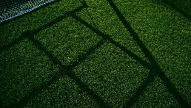 Diagonal goalpost shadow creating rectangular grid across sunlit soccer grass pitch