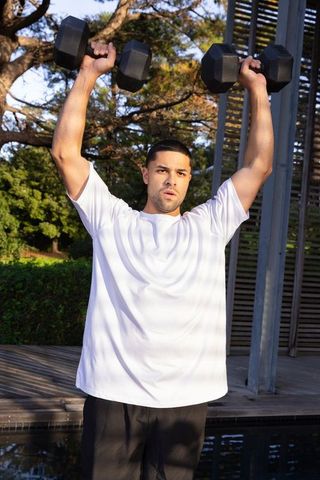 A man raises hexagonal dumbbells overhead in an outdoor setting beside reflective water, exemplifying an active and healthy lifestyle. Ideal for fitness promotions, wellness blogs, strength training programs, or advertisements focusing on healthy living and exercising in natural environments. The image's composition highlights outdoor physical activity and promotes the concept of incorporating nature into a workout routine.