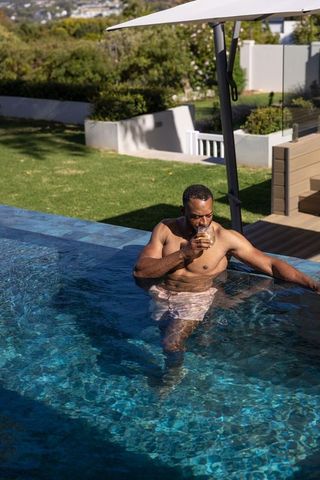 Mature african man relaxing in pool sipping drink by luxury backyard deck and umbrella