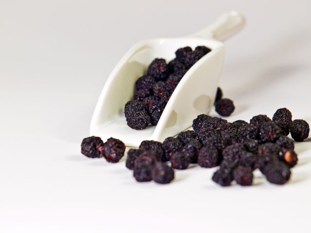 Dried aronia berries spilling from a white ceramic scoop on a white background. This image highlights the importance of healthy snacks and the benefits of antioxidants. Perfect for articles on superfoods, recipes, health blogs, or promotional materials for healthy eating and organic food stores.