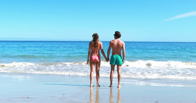 Two people, holding hands, are enjoying a sunny day at the ocean beach, gazing at the horizon and standing on the shoreline. This could be used for travel brochures, romance-themed materials, summer vacation promotions, or any content related to coastal destinations and outdoor activities.
