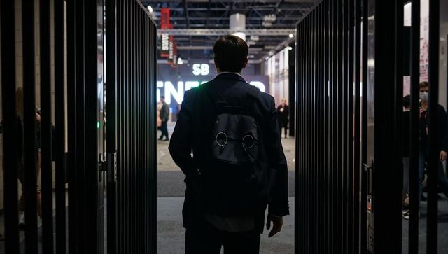 Asian man walking through metal gates into expo hall wearing blazer and ringed black backpack