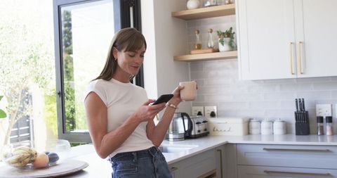 Middle-aged Woman Enjoying Coffee and Smartphone in Modern Kitchen