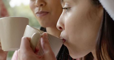 Asian women sipping from beige ribbed mugs by window, cozy morning routine