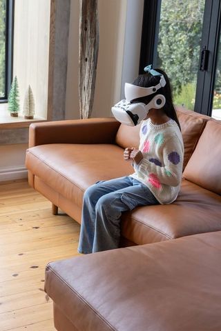 Young asian girl wearing white vr headset sitting on leather sofa experiencing virtual reality
