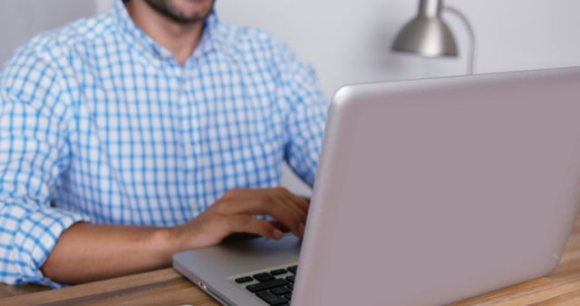 Man wearing a blue checkered shirt working on laptop at wooden desk in modern office. Ideal for illustrations of remote work, freelancing, business tasks, productivity in a modern workspace, professional tech use, and casual work environments.