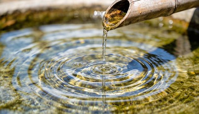 Bamboo fountain pouring clear water droplet into stone basin creating concentric ripples