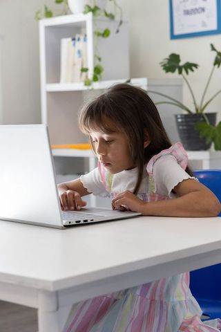 Young girl concentrating on laptop while learning at home in pastel dress and workspace