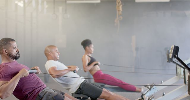 Three diverse individuals exercising on rowing machines in an indoor gym. They are focused and determined, engaging in a full-body workout. Suitable for fitness, healthy living, gym promotions, personal training services, and athletic training content.