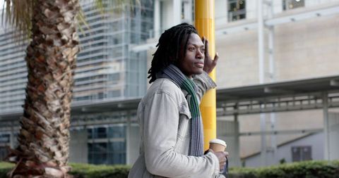 Contemplative african american man leaning on yellow pole holding coffee in modern city