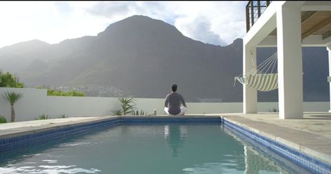 Indian man meditating on blue-tiled pool terrace facing mountain view, holding cup, calm