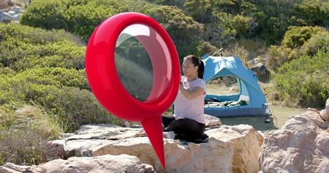 Woman packing camping gear on sunlit coastal rocks with blue tent and red location marker