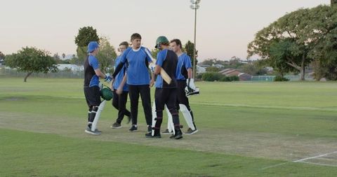 Male cricket teammates chatting and exchanging gear on pitch before match at dusk