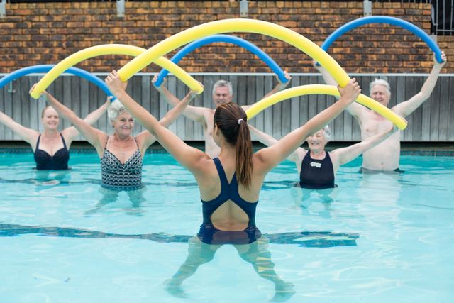 Group of senior swimmers participating in a water exercise class, led by a female instructor. They are using pool noodles for resistance training in a swimming pool. Ideal for promoting senior fitness, aquatic exercise programs, and healthy lifestyle activities for older adults.